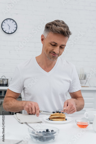 Positive man cutting pancakes near blurred blueberries and honey in kitchen.