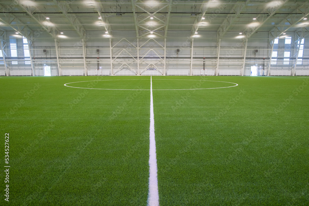 Fototapeta premium A new empty European sports hall for playing football at any time of the year. The football field is covered with artificial grass, lighting spotlights are installed on the walls and ceiling.