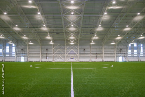 A new empty European sports hall for playing football at any time of the year. The football field is covered with artificial grass, lighting spotlights are installed on the walls and ceiling.