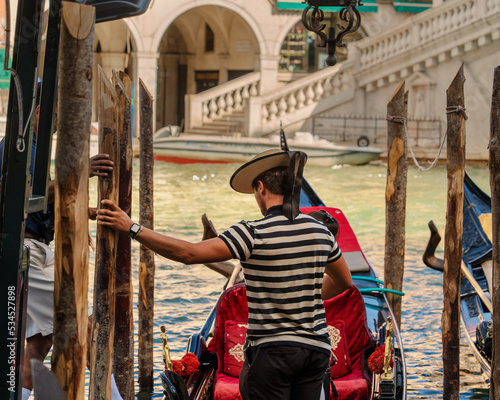 The gondolier helps tourists enter the gondola near the bank of the canal of the city of Venice on a sunny morning, the Venetian gondolier extended a helping hand to the passengers of the boat