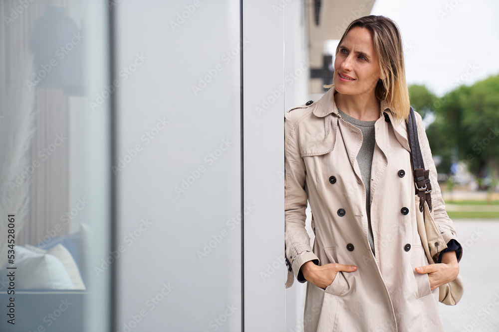 Mid-shot of woman wearing raincoat looking at shop window while leaning on a wall Mid-shot of woman wearing raincoat looking at shop window while leaning on a wall