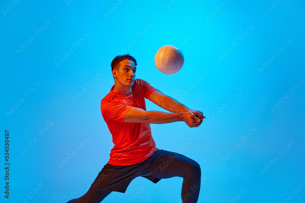 One male volleyball player in orange-black uniform training with ball isolated on blue studio background in neon light. Sport, gym, team sport, challenges