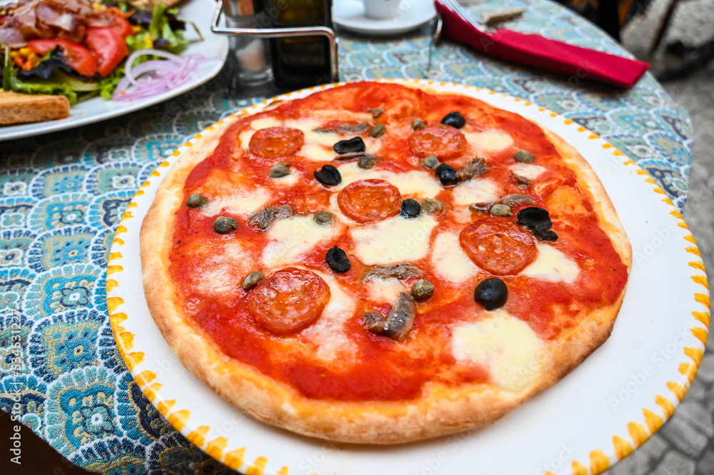 Pizza on the table in restaurant in Italy. Spicy sausage, vegetable, olives and cheese on pizza served on plate. 