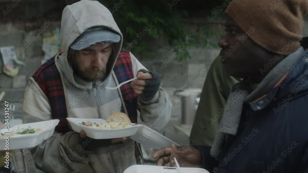 Group of multiethnic homeless men talking while eating free meal served by volunteers on outdoor soup kitchen Group of multiethnic homeless men talking while eating free meal served by volunteers on outdoor soup kitchen
