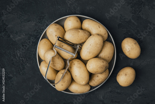 Potato. Fresh raw organic potatoes in bowl with peeler on dark stone background. photo