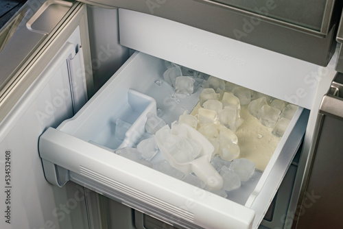 Inside of automatic ice maker with a drawer of ice cubes and plastic scoop. High angle view, closeup pile of ice cubes in a drawer of automatic ice maker in modern refrigerator.