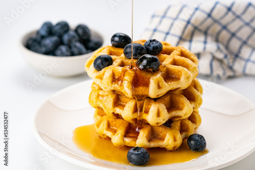 Closeup view of pouring honey on stack of fresh delicious homemade waffles topping with ripe organic blueberry fruit in white ceramic dish.