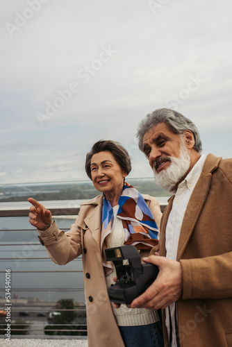 happy senior man holding vintage camera near wife smiling and pointing with finger on bridge.