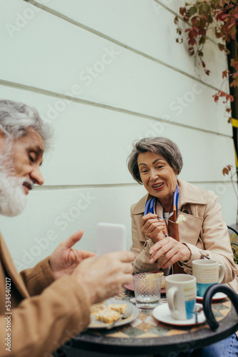 bearded senior man showing smartphone to cheerful wife during breakfast on terrace of cafe.