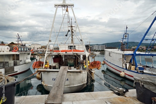 White fishing boat moored in port in town of Koper near the city center. Around it are other fishing vessels. In horizon is landscape with hills and overcast sky.