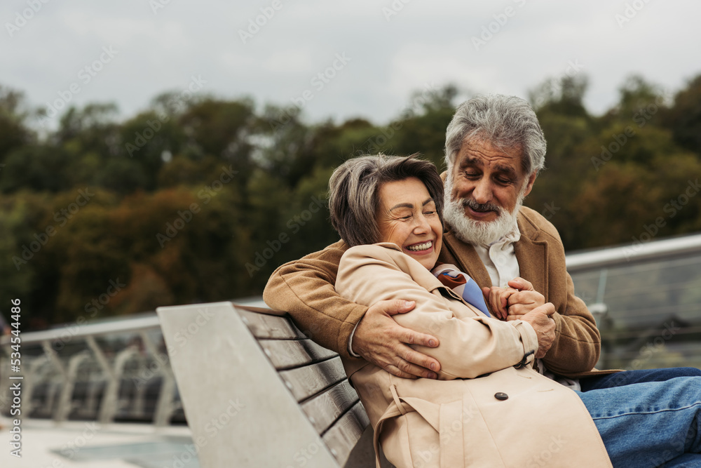 Fototapeta premium happy senior man hugging joyful wife while sitting on bench outdoors.