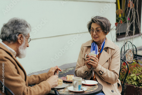 senior woman in eyeglasses and trench coat holding cup during brunch with husband.
