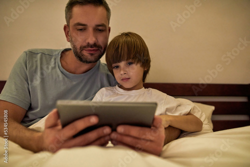 Dad and son watch digital tablet in bed at evening