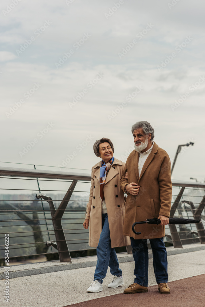 full length of cheerful senior woman walking with bearded husband holding umbrella near guard rail of bridge. full length of cheerful senior woman walking with bearded husband holding umbrella near guard rail of bridge.
