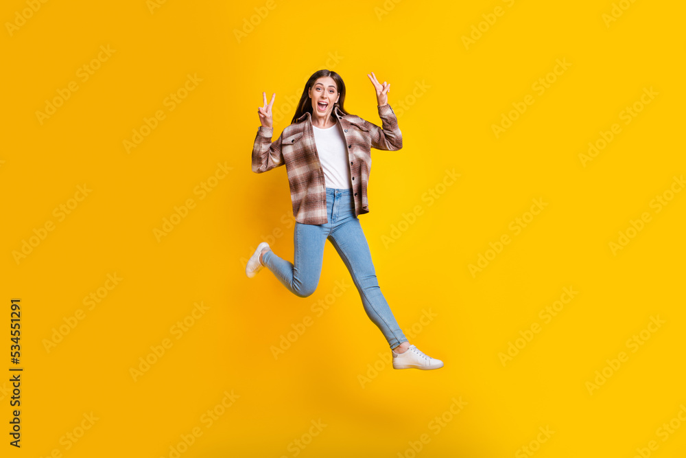 Full length photo of pretty shiny girl dressed plaid shirt jumping high showing v-signs isolated yellow color background