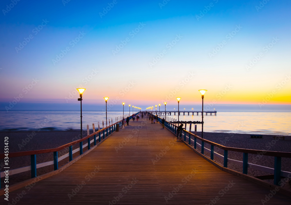 Illuminated romantic vintage Palanga pier after sunset with people walking in beautiful summer evening. Lithuania summer holiday landmarks destination