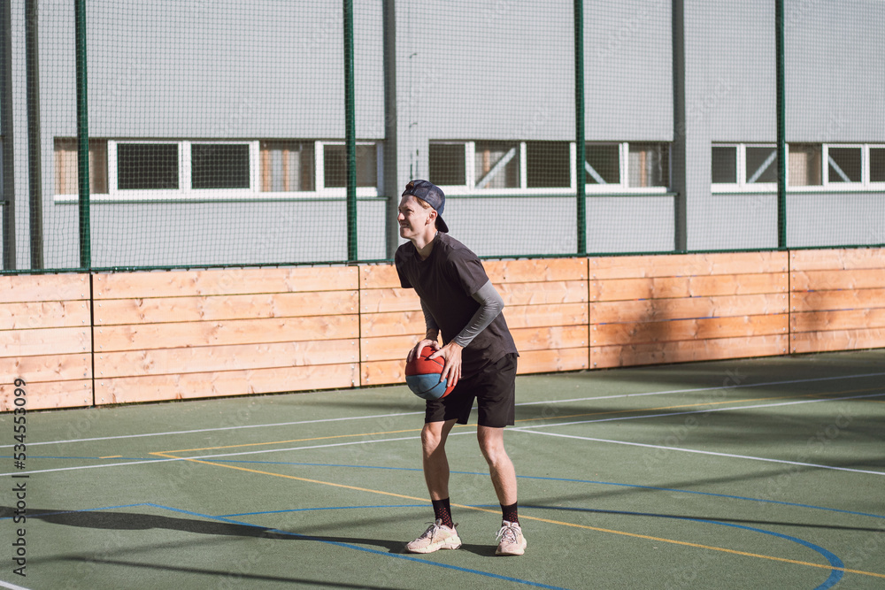 Blond boy in sportswear practices shooting a basketball from behind the three-point line. Outdoor basketball court. Preparing for the season Blond boy in sportswear practices shooting a basketball from behind the three-point line. Outdoor basketball court. Preparing for the season