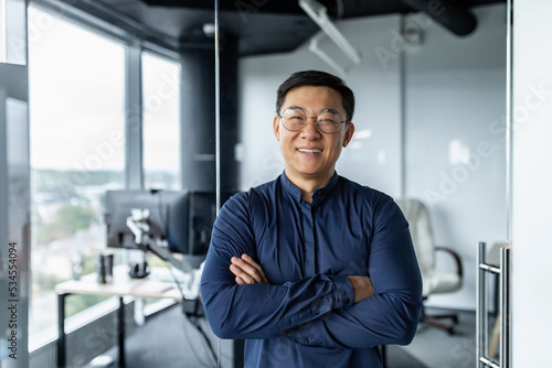 Portrait of successful asian businessman with crossed arms, businessman investor working inside office building loft, looking at camera and smiling, satisfied investor financier. © Liubomir