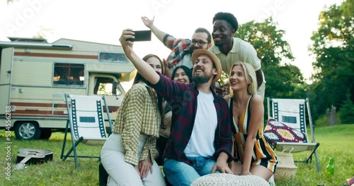 A group of friends takes a selfie as a souvenir for a scrapbook of their trip together. Students fool around making funny faces for their phones. In the background outdoor park camper.