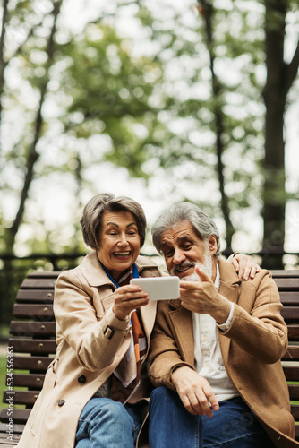 senior woman smiling while holding smartphone near bearded husband in coat.