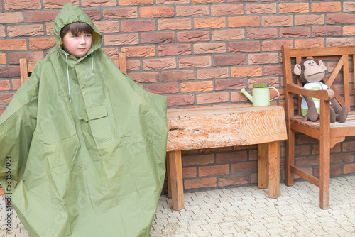 Boy in the green raincoat is sitting and waiting for raining stop. Fall rainy weather season. Brick wall background. Wooden chair and tale. Child want to go outside. photo