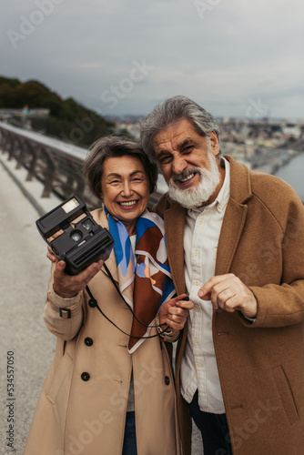 cheerful senior woman in trench coat holding vintage camera near happy husband outside.