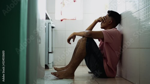 Depressed hispanic young man sitting on kitchen floor covering face in shame. Desperate South American person during difficult hard times suffering from mental illness photo
