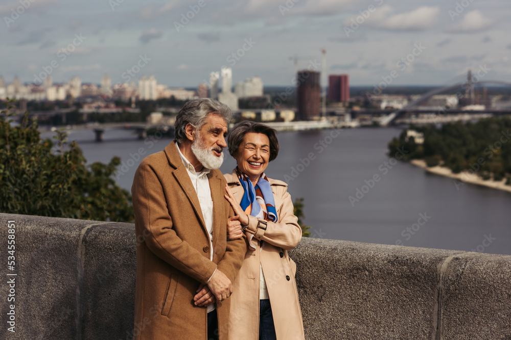 happy senior couple in beige coats smiling and walking on bridge near river.