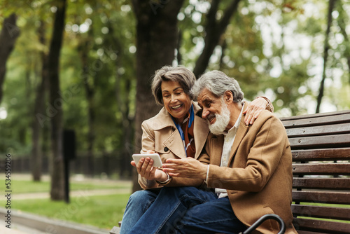 happy senior couple in coats sitting on bench and looking at smartphone.