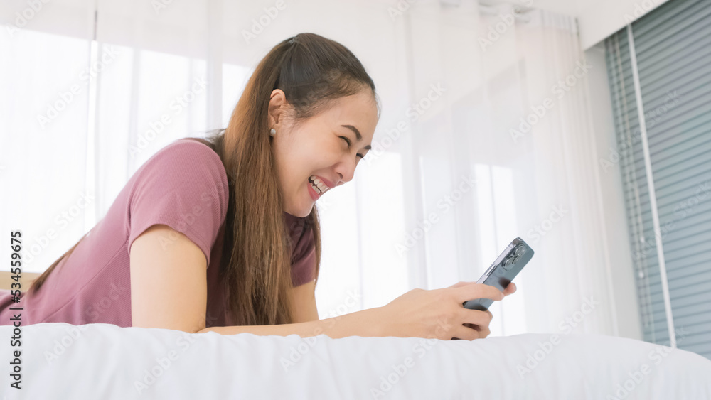 Happy Asian woman laying in the bed using a smartphone with a smile and laughing in the morning. Women use device for social networks, chat or online shopping from smartphones. Happy Asian woman laying in the bed using a smartphone with a smile and laughing in the morning. Women use device for social networks, chat or online shopping from smartphones.