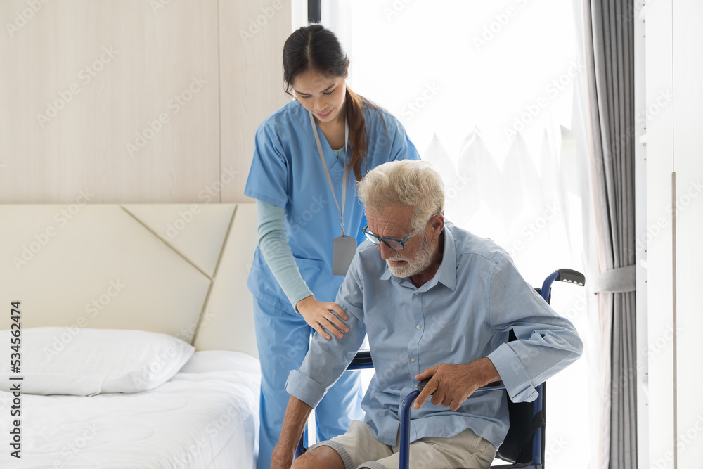 Fototapeta premium Young asian female nurse and patient senior man. Female nurse caring old man walk from wheelchair at hospital ward. Nurse helping elderly man walk from wheelchair at nursing home