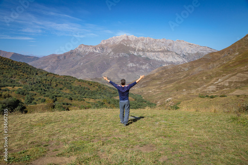 An adult man stands on top of a mountain with outstretched arms.