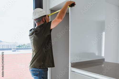 Un hombre con casco de obra mide con un metro el hueco de un mueble. Montador de muebles. Instalación de cocinas. Hueco para una nevera.
