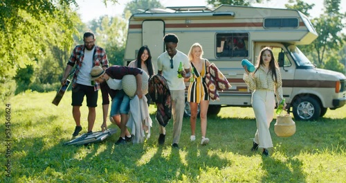 Friends arrive for an open-air camper trip to the lake together. They organize a picnic spread out deck chairs poufs for relaxing and barbecuing.