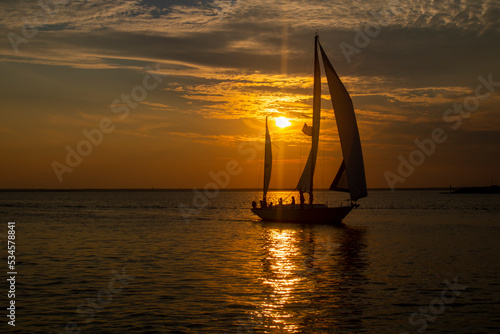 sail boat in ocean waters on a beautiful yellow Orange brown sunrise sunset with sun behind it's sails
