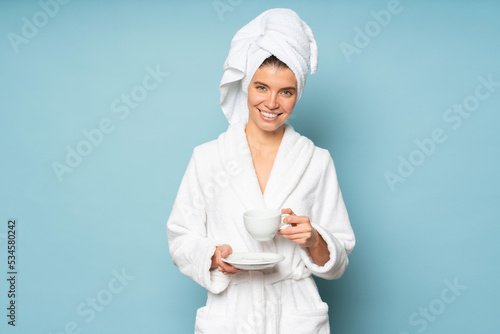 Portrait of woman in bathrobe and towel on head standing on blue background with cup of coffee photo