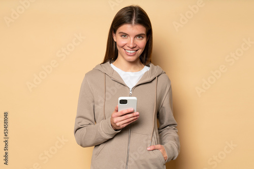 Woman in hoodie downloading fitness app to her phone to train in morning isolated on brown