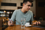 A young guy using his computer in cafe and talking on the phone while drinking water and coffee 