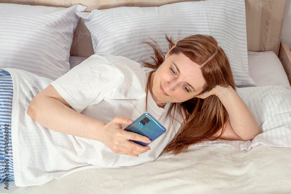 Woman with smartphone in bed at home or hotel in the morning. Lady relaxing in a nightgown. Girl wearing nightie on clean white bedding, cozy blanket Woman with smartphone in bed at home or hotel in the morning. Lady relaxing in a nightgown. Girl wearing nightie on clean white bedding, cozy blanket
