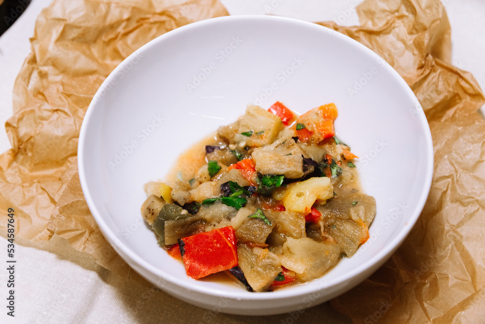 Eggplant stew with vegetables. Large cubes of stewed vegetables in a deep white ceramic bowl. View from slightly above. The dish is served hot. Ingredients: cauliflower, red bell peppers, parsley. Eggplant stew with vegetables. Large cubes of stewed vegetables in a deep white ceramic bowl. View from slightly above. The dish is served hot. Ingredients: cauliflower, red bell peppers, parsley.
