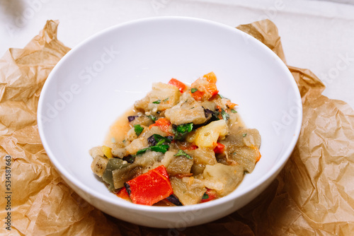 Eggplant stew with vegetables. Large cubes of stewed vegetables in a deep white ceramic bowl. View from slightly above. The dish is served hot. Ingredients: cauliflower, red bell peppers, parsley.