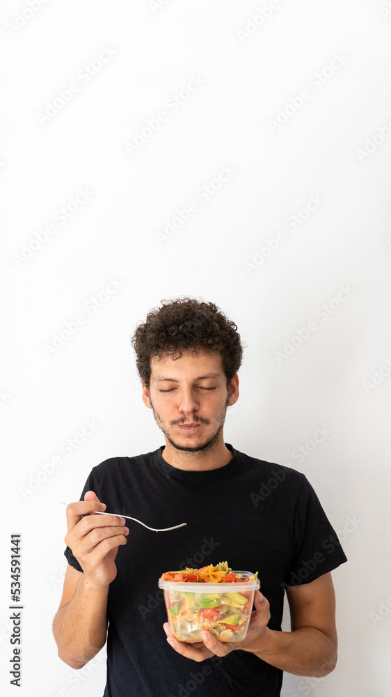 Happy young caucasian man eating and holding a healthy mediterranean pasta salad tupperware Happy young caucasian man eating and holding a healthy mediterranean pasta salad tupperware