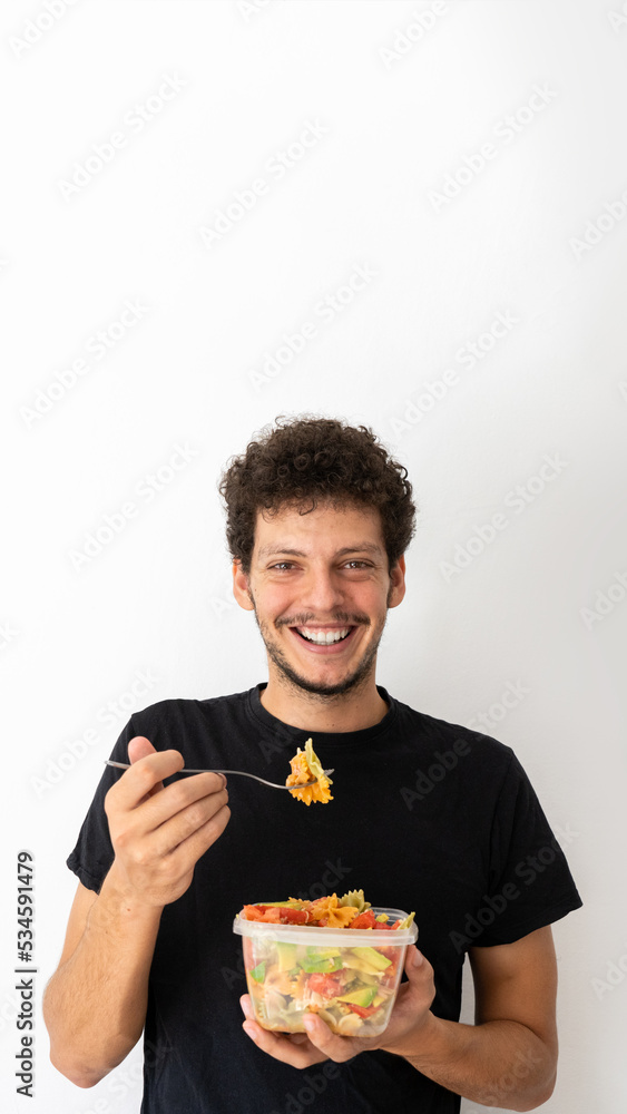 Happy young caucasian man eating and holding a healthy mediterranean pasta salad tupperware Happy young caucasian man eating and holding a healthy mediterranean pasta salad tupperware