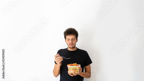 Happy young caucasian man eating and holding a healthy mediterranean pasta salad tupperware