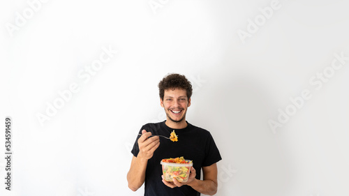 Happy young caucasian man eating and holding a healthy mediterranean pasta salad tupperware