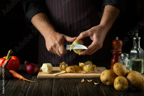 Chef peels raw potatoes in a hotel kitchen before preparing a national dish. Close-up of a cook hands with a knife while working.