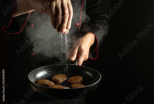Fototapeta Naklejka Na Ścianę i Meble -  Professional chef adds salt to cutlets that are fried in a pan. Cooking meat patties for a burger. Menu for a restaurant or hotel on a black background