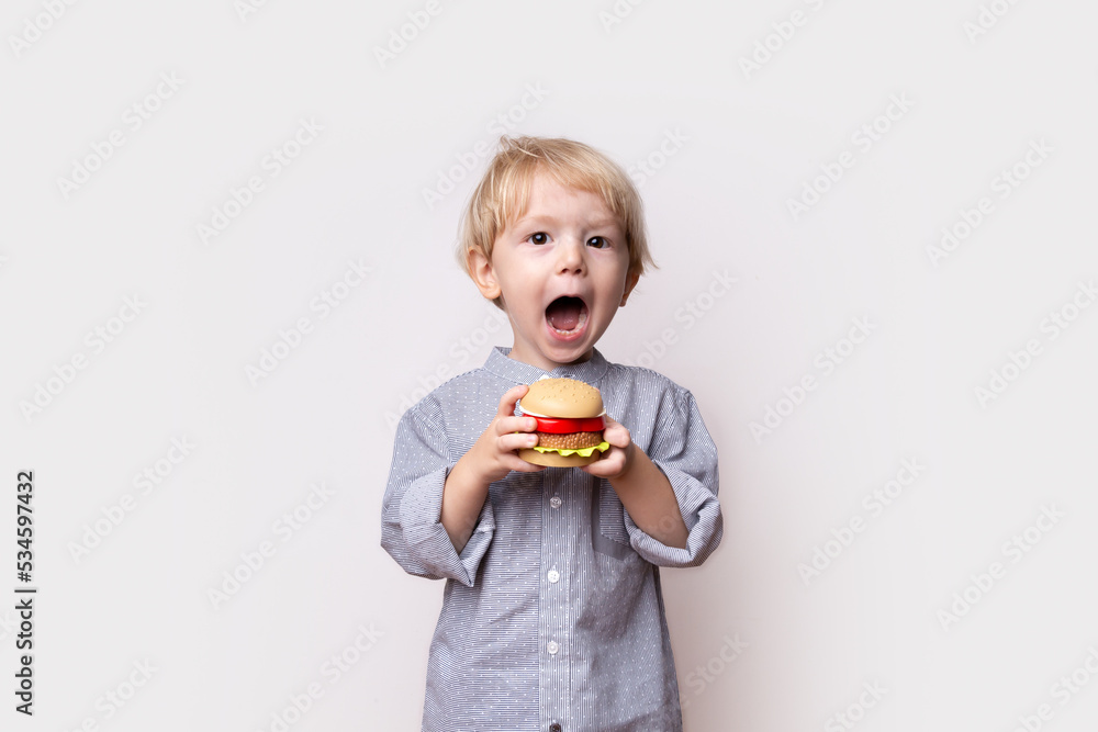 little toddler boy with blond hair in a shirt holds a toy plastic hamburger in his hands on a white background, feeding children, hungry child, food in kindergarten, healthy eating, space for text little toddler boy with blond hair in a shirt holds a toy plastic hamburger in his hands on a white background, feeding children, hungry child, food in kindergarten, healthy eating, space for text