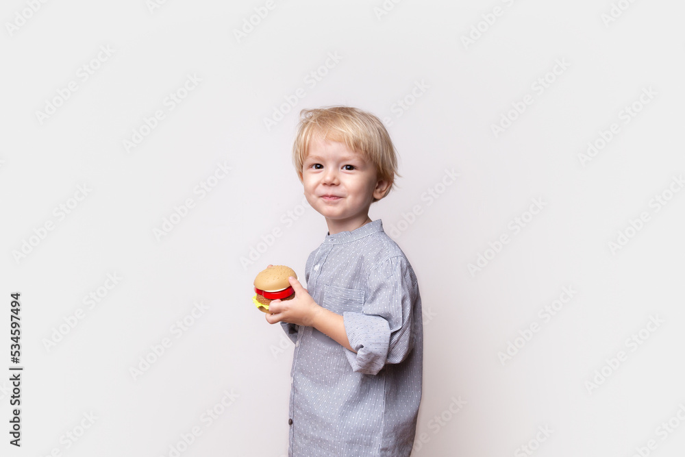 little toddler boy with blond hair in a shirt holds a toy plastic hamburger in his hands on a white background, feeding children, hungry child, food in kindergarten, healthy eating, space for text little toddler boy with blond hair in a shirt holds a toy plastic hamburger in his hands on a white background, feeding children, hungry child, food in kindergarten, healthy eating, space for text