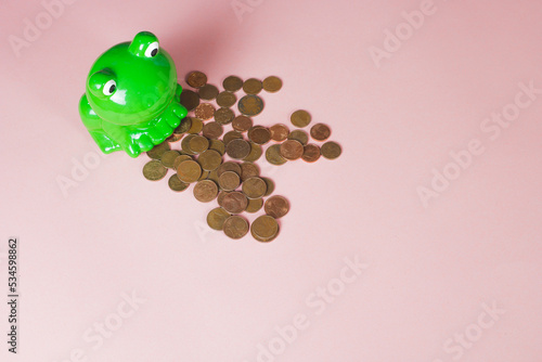 Ceramic moneybox, shape and color of green frog. Euro coins poured out from the cash bank on the pink background with copy space for text. Concept of spending savings. © Matej